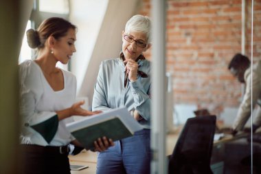 Mature CEO talking to young colleague who is showing her business reports in the office.