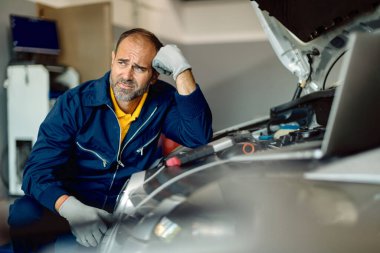 Pensive auto repairman feeling worried while examining car engine in a workshop. 