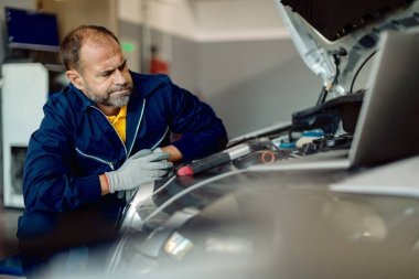 Worried mechanic examining car engine while working in auto repair shop. 