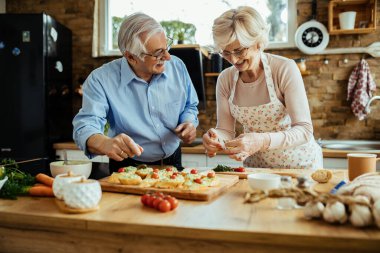 Happy mature couple talking and having fun while making bruschetta together in the kitchen. 