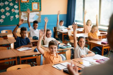 Large group of happy school kids raising their arms to answer teacher's question on a class in the classroom. 