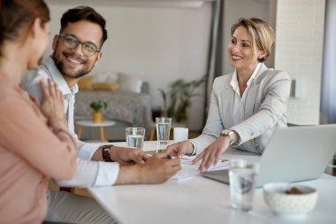 Happy real estate agent closing a deal with young couple and offering them a contract to sign during a meeting. 