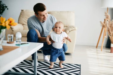 Cute baby boy learning to walk with help of his father at home. 