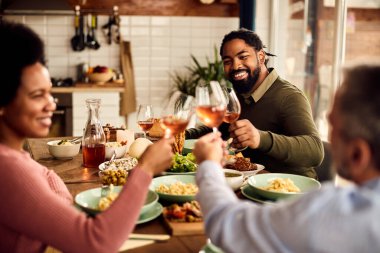 Group of happy friends toasting with wine while having meal together at dining table. Focus is on black man. 