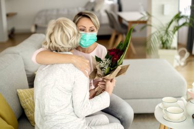 Happy woman with face mask embracing senior mother while bringing flowers and visiting her at home during coronavirus epidemic. 