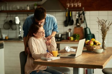 Caring man kissing his girlfriend while she is using computer and working at home.