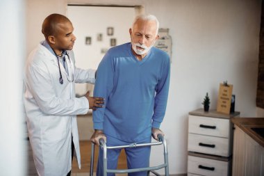 African American doctor assisting senior man in walking with mobility walker during physical therapy at nursing home. 