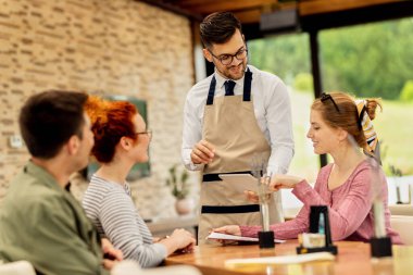 Smiling waiter showing menu on digital tablet to happy woman who came with friends in a cafe. 