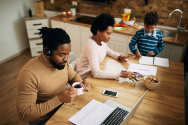 Black father drinking tea while analyzing business reports and using laptop at home. 