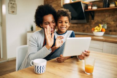 Happy black mother and son using touchpad and greeting someone while having video call at home. 