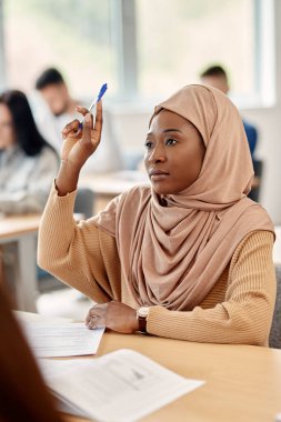 Muslim college student raising her hand to answer a question while  attending a class in lecture hall.