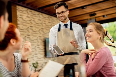 Happy waiter using touchpad while communicating with guests in a restaurant. 