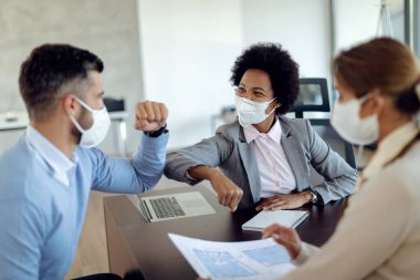 Happy African American real estate agent and her clients greeting with elbows in the office. They are wearing protective face masks due to COVID-19 epidemic. 