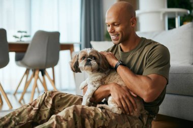 Genç Afrikalı Amerikalı asker evde birlikte vakit geçirirken köpeğine sarılıyor..