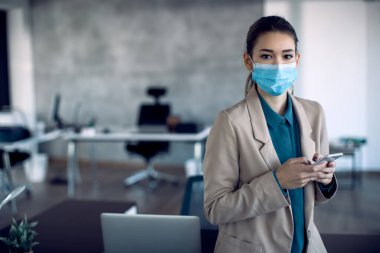 Young businesswoman with face mask texting on mobile phone in the office and looking at camera.