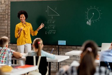 Happy black elementary school teacher talking to her students and explaining them mathematics on blackboard in the classroom. 