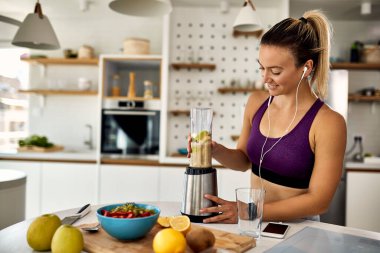 Young happy sportswoman using blender and making fruit smoothie in the kitchen. 