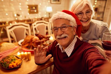 Happy senior man and his wife waving to camera while taking selfie on Christmas Eve at dining room. 