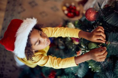 High angle view of small black girl decorating Christmas tree at home. 