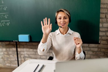 Happy teacher greeting her students and waving while holding online class from the classroom. 