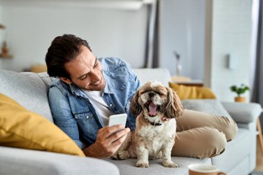 Happy man using smart phone while relaxing on the sofa with his dog who is yawning. 