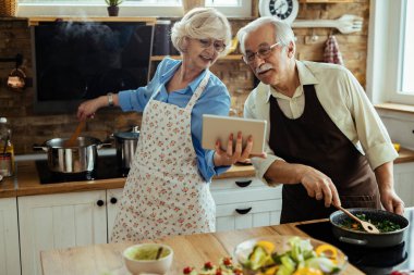 Senior husband and wife using digital tablet while cooking together in the kitchen. 