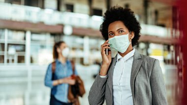 Virüs salgını sırasında havaalanında yüz maskesi takıp akıllı telefondan konuşan siyahi kadın yolcu..