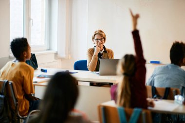 Mature teacher asking a question during a class while one of her students is raising hand to answer it at elementary school.