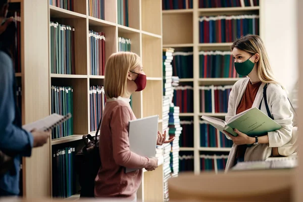 Female students with face masks communicating while wearing protective face masks at college library.