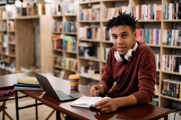 Young smiling black student using laptop and studying in library while looking at camera.