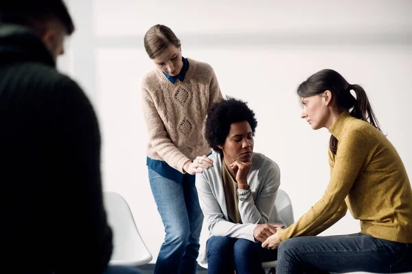 Sad black woman getting support from members of group therapy at community center.