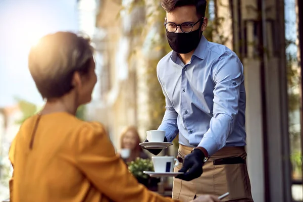 Young waiter wearing protective face mask while serving coffee to a customer in a cafe after reopening.