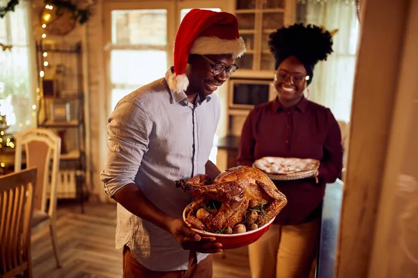 Young black couple serving food for Christmas lunch at home. Man is serving roast turkey while woman is bringing the pie.