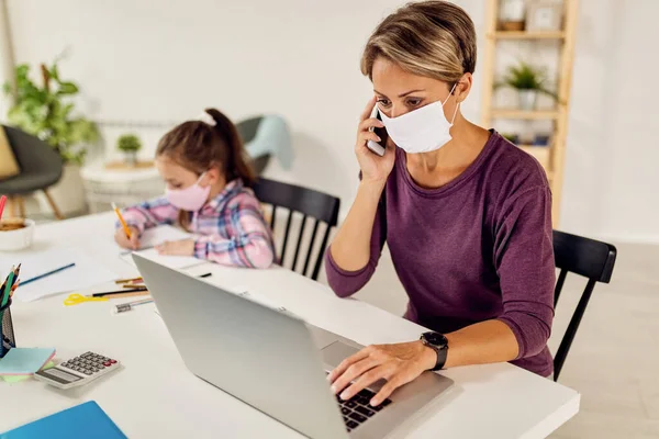 Working mother using laptop and talking on the phone while her daughter is studying at home during coronavirus pandemic.