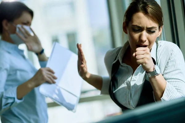 Young businesswoman coughing into elbow while being with her colleague in the office.