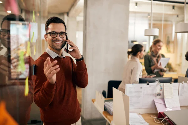 Happy entrepreneur examining mind map on glass wall while communicating on cell phone at the office.