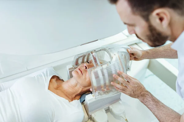 Medical technician preparing senior patient for head MRI scan in examination room at clinic. Focus is on woman.