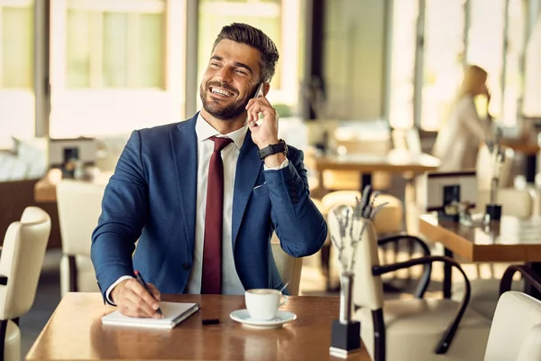 Cheerful businessman taking notes while making a phone call over cell phone in a cafe.