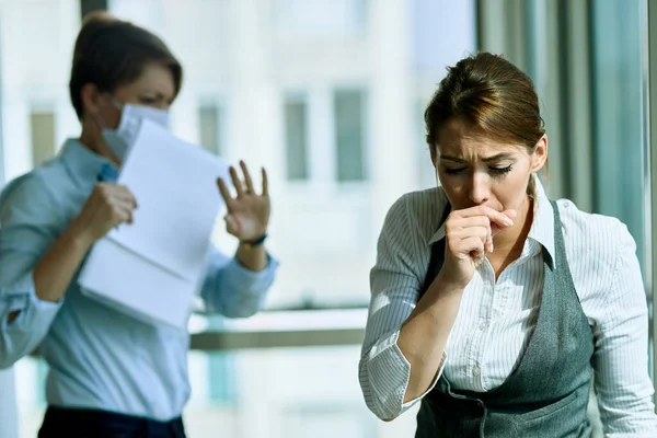 Young businesswoman feeling sick and coughing while being in the office. Her colleague is in the background.
