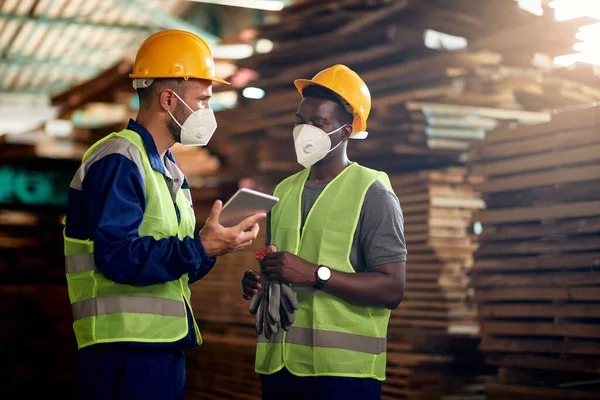 Manual workers wearing face masks while talking and using touchpad in wood warehouse.
