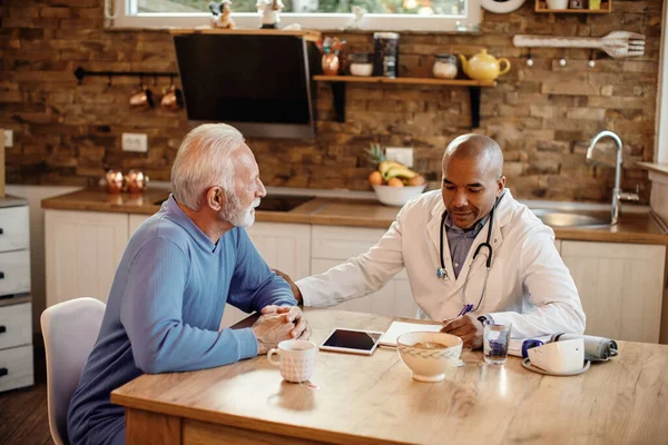 Happy black doctor taking notes while talking to senior man during home visit. 