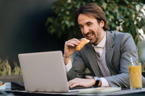 Young happy businessman eating croissant while surfing the net on laptop in outdoor cafe.