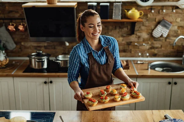 Young smiling woman holding cutting board with healthy bruschetta she has prepared in the kitchen.