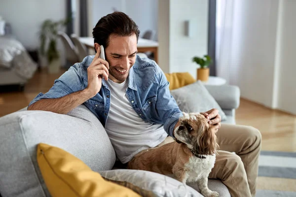 Happy man talking on mobile phone while enjoying with his dog on the sofa.