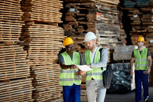 Warehouse manager and African American worker going through checklist at lumber distribution department.