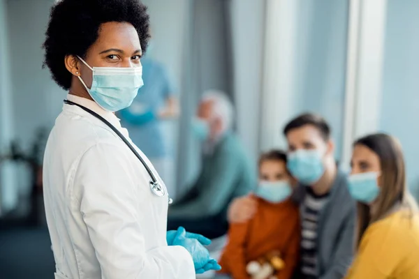 Black female doctor with face mask standing in a hallway and looking at camera. Her patients are sitting in the background.