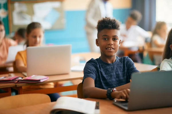 Smiling African American schoolboy e-learning on laptop during a class at elementary school and looking at camera.