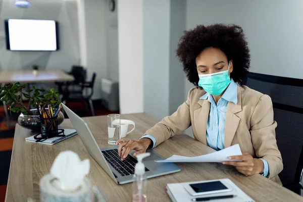 African American female entrepreneur analyzing business reports and working on a computer while wearing face mask due to coronavirus pandemic.
