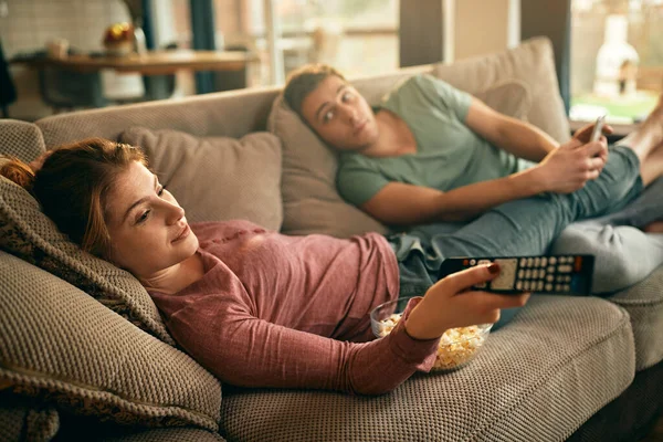 Smiling woman relaxing with her boyfriend on the sofa and changing channels on TV at home.