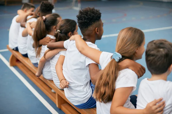 Back view of multi-ethnic group of elementary students embracing while sitting on a bench during physical activity class at school gym.